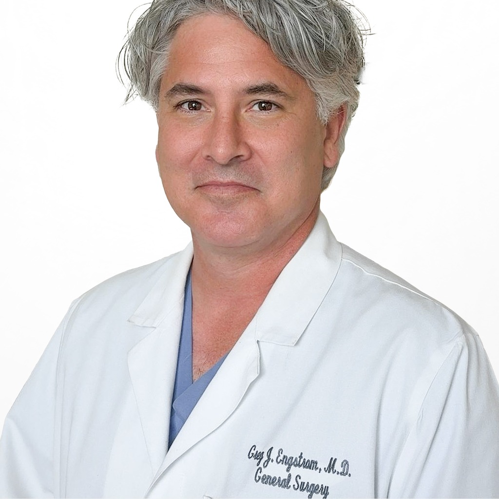 13. Surgeon. Middle-aged male doctor in a white lab coat and blue scrubs against a solid white background. Used by most academic hospitals