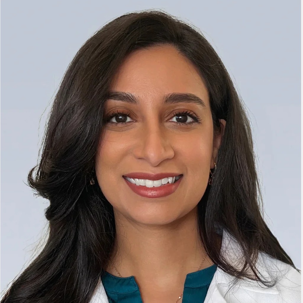 15. Female cardiologist. Smiling woman in a white lab coat over a teal top, set against a plain light gray background. Coat plus a clean background