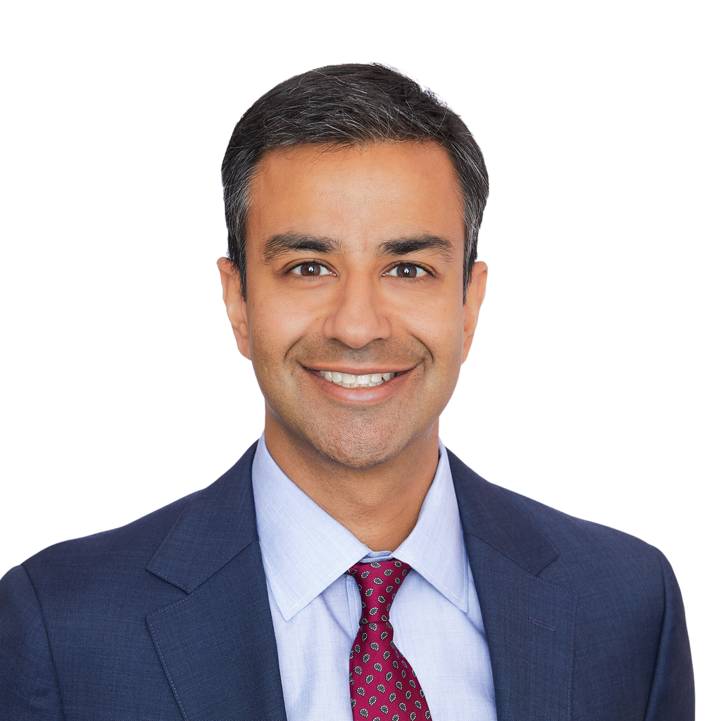 22. Financial analyst. Smiling South Asian man in a dark suit and red tie, set against a solid black background. Junior staff. Tie still on