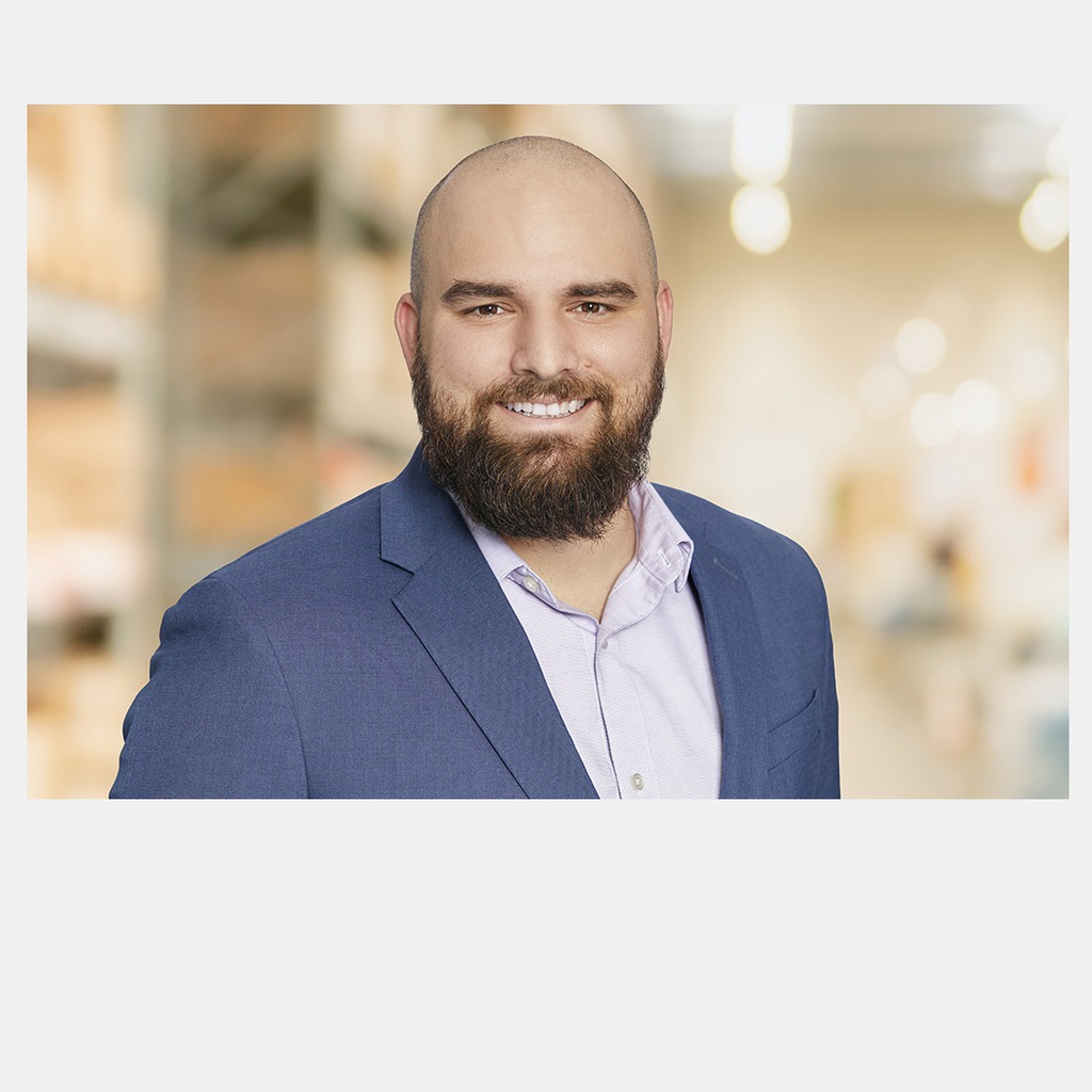 26. Bank branch manager. Smiling man with a beard, wearing a blue blazer, against a blurred office background. Retail banking softens the edges