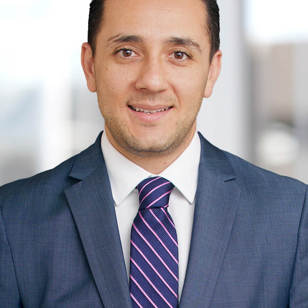 31. General counsel. Man in a blue suit and striped tie, smiling with his hands in his pockets, against a blurred modern office background